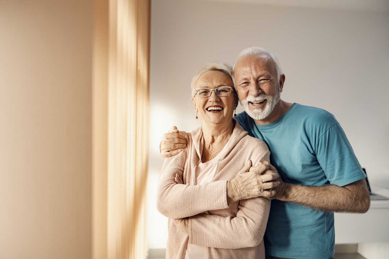 Senior couple hugging in a nursing home. A happy senior couple standing next to a window in a nursing home, hugging and smiling. They have all care they need. Exceptional memory care in Springfield Oregon