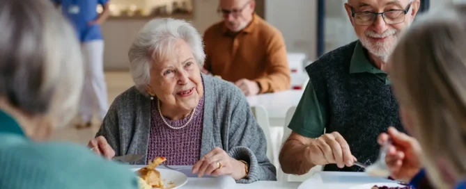 Seniors sharing lunch in a community dining room, showing assisted care vs in-home care and daily social support.
